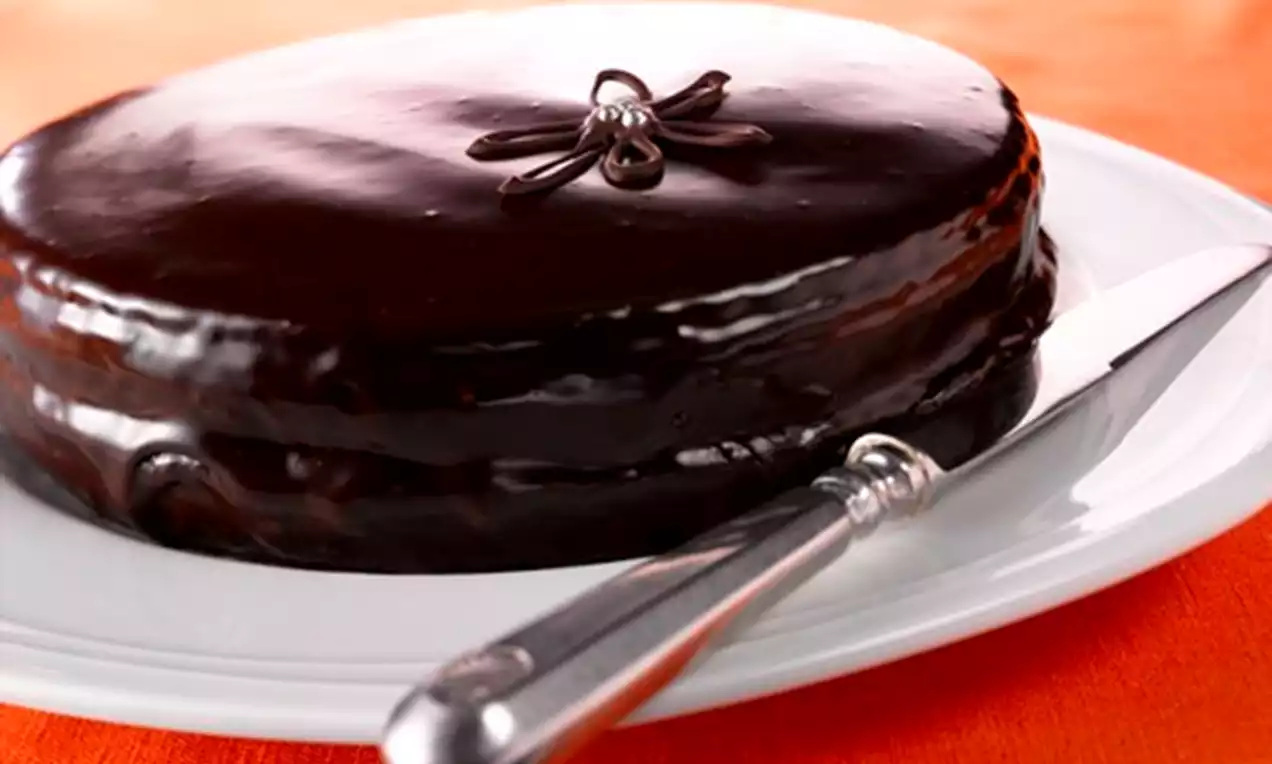 Pyöreä sacherkakku, jonka kiiltävä suklaakuorrute peittää tasaisesti, pieni koriste päällä valkoisella lautasella hopeisella kakkulapiolla