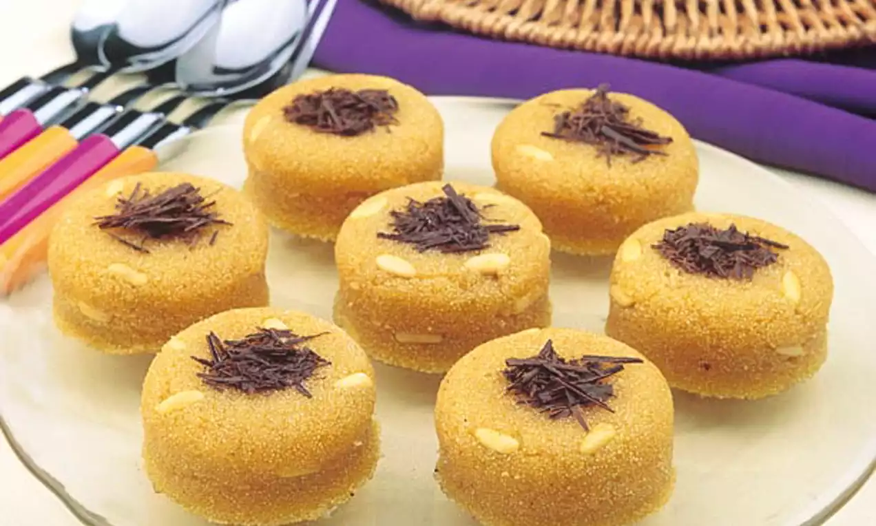 Altın rengi yuvarlak irmik helvaları, üzerlerinde ince çikolata parçaları ve içlerinde çam fıstığıyla cam bir tabakta düzenli şekilde sunulmuş