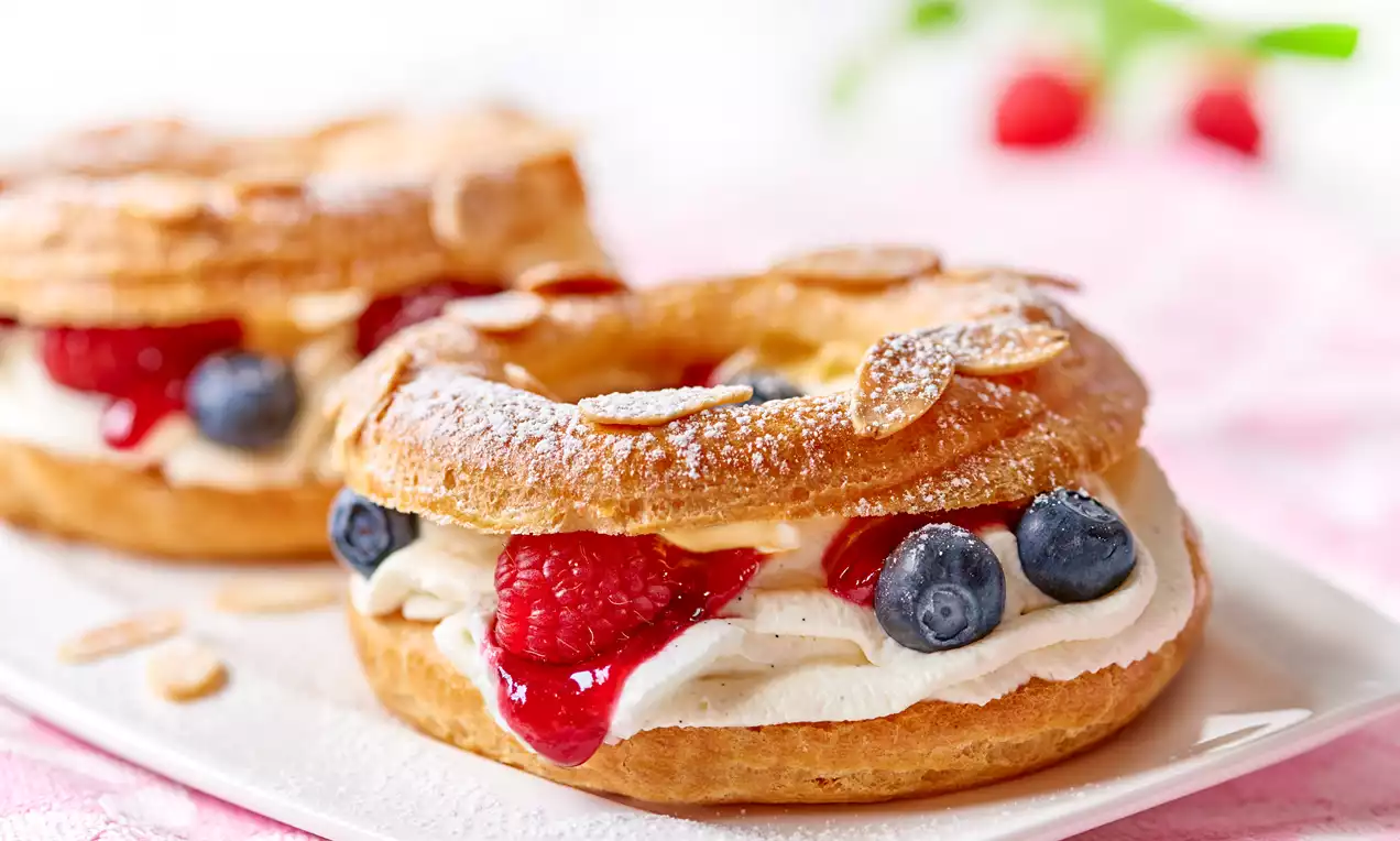 Złociste ciasto parzone w kształcie pierścienia posypane migdałami, przełożone kremem waniliowym, malinami, borówkami i sosem owocowym na białym talerzu