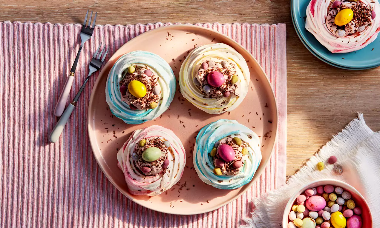 Colourful meringue nests swirled with pastel pink and blue, filled with chocolate shavings and speckled eggs on a peach plate