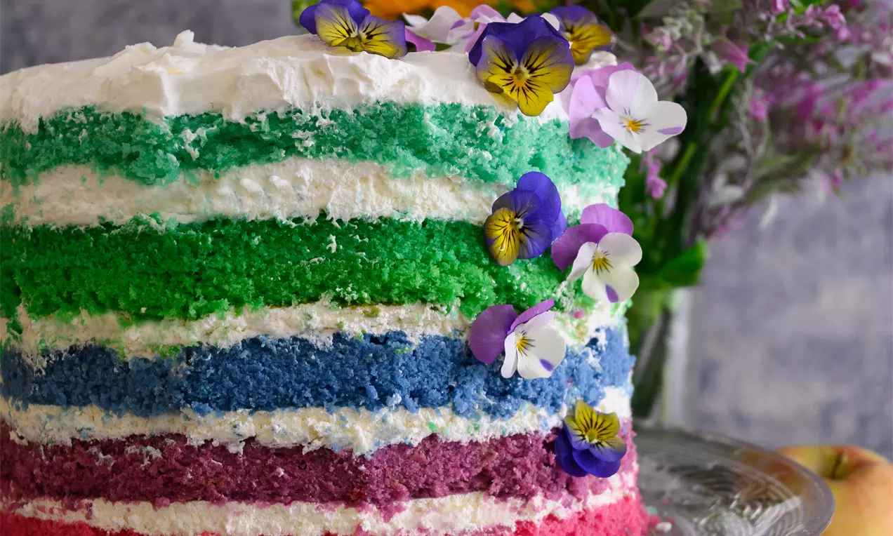 Висока торта с цветни пластове в розово, лилаво, синьо и зелено, редувани с пухкави бели кремове, украсена с цветя върху стъклена чиния