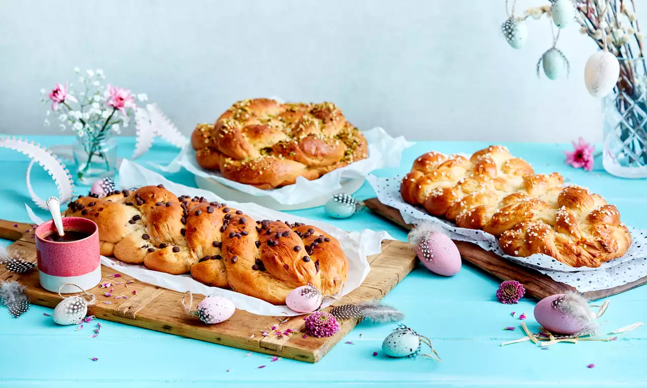 Braided Easter breads golden brown with glossy crusts, topped with chocolate chips, nuts, or pearl sugar on wooden boards with pastel eggs