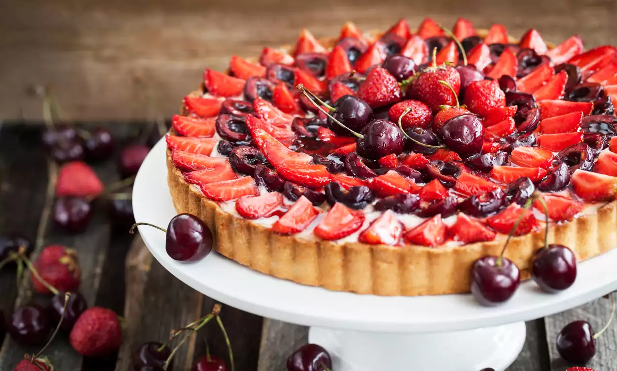 Fruittaart met een knapperige lichtbruine korst, bedekt met glanzende rode aardbeien, kersen en donkere bessen op een wit taartplateau