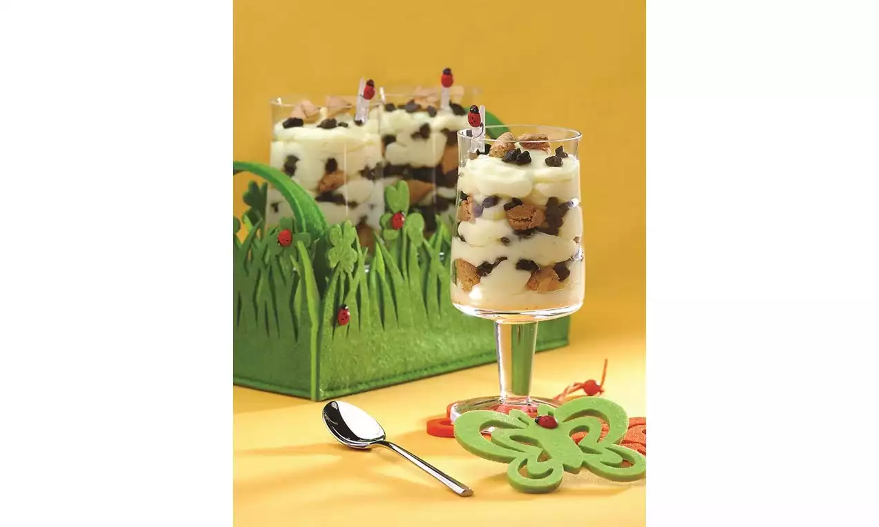 Uzun ayaklı cam bardakta vanilya kreması ve kek parçaları katmanlı, kelebek şeklinde yeşil süsleme ve çim desenli arka plan detaylı