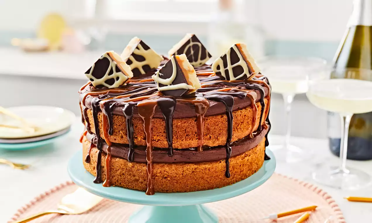 Gluten-free billionaire's cake with golden sponge layers, glossy chocolate and caramel drips, topped with chocolate-topped caramel cubes