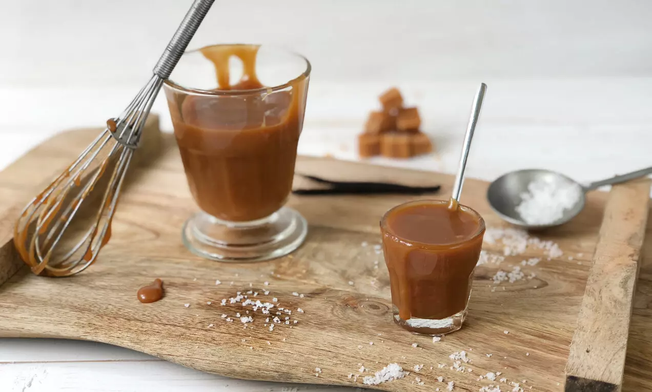 Гладък светлокарамелен сос с лъскава кремообразна текстура, поднесен в стъклени чаши върху дървена дъска с щипки морска сол