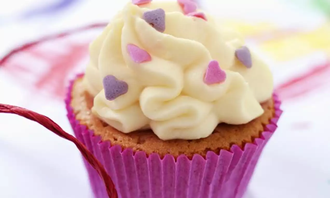 Vanillekleurige slagroomtoef op een goudbruine cupcake in een roze papiertje, versierd met paarse en roze hartjesconfetti