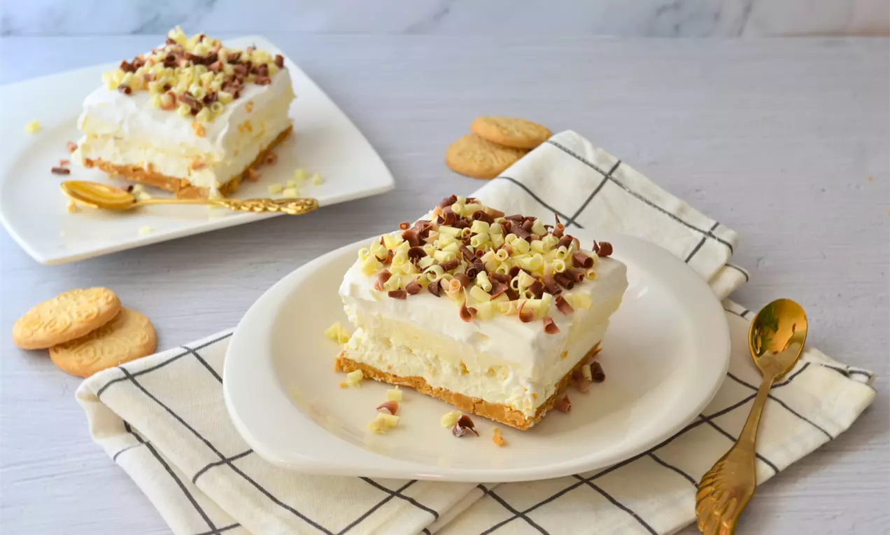 Светла квадратна торта с пухкава бяла пълнеж върху златист бисквитен блат, поръсена с фини къдри от бял и млечен шоколад върху чиния