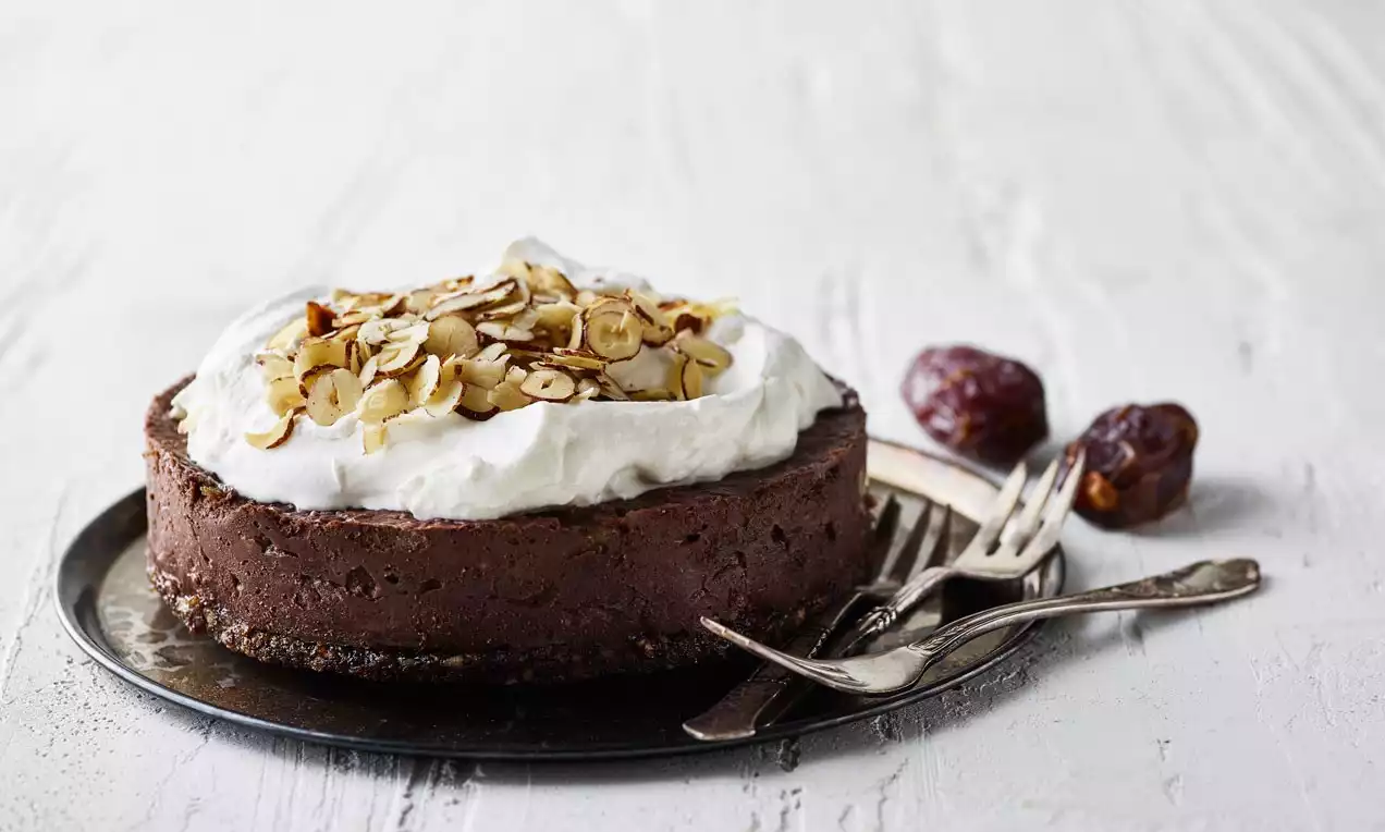 Mörkbrun vegansk chokladmoussekaka med vit gräddtopping och rostade nötflingor på svart tallrik, intill bestick och dadlar på ljus yta