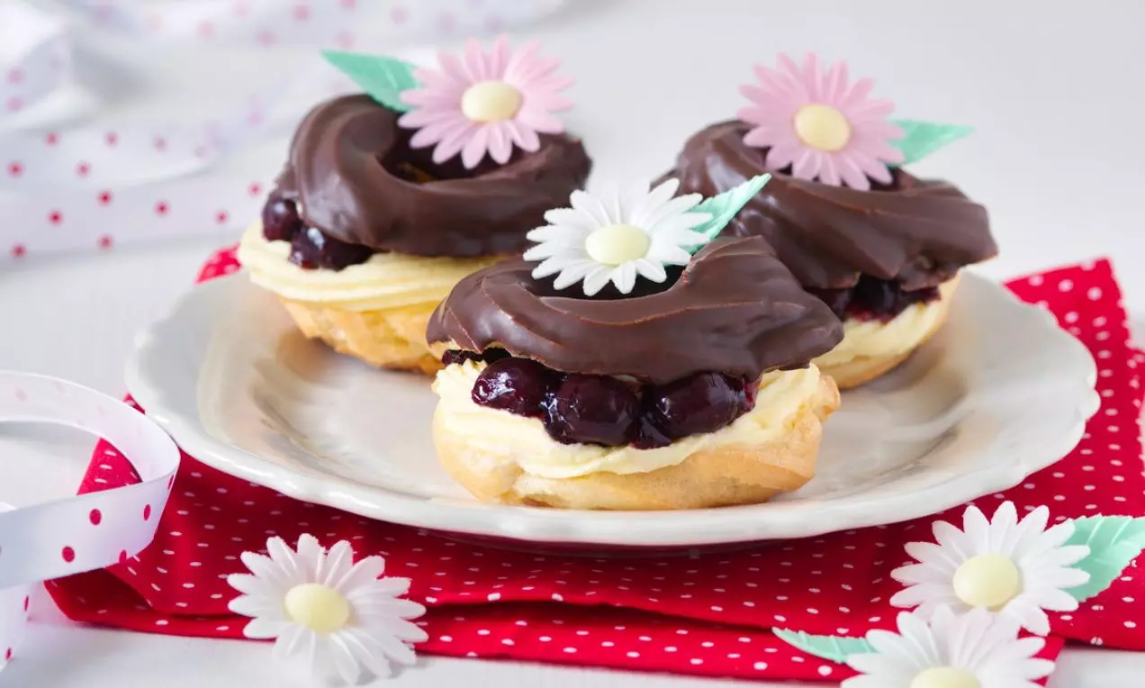 Na bílém talíři leží světlé odpalované těsto s lesklým tmavým čokoládovým krémem, zdobené růžovými a bílými květinkami na červeném ubrousku