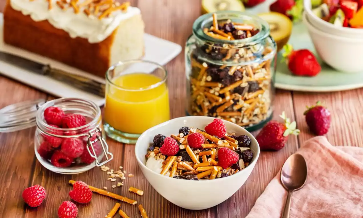 Granola doré mêlé de sticks croquants, fruits secs et framboises fraîches dans un bol blanc sur une table en bois éclairée