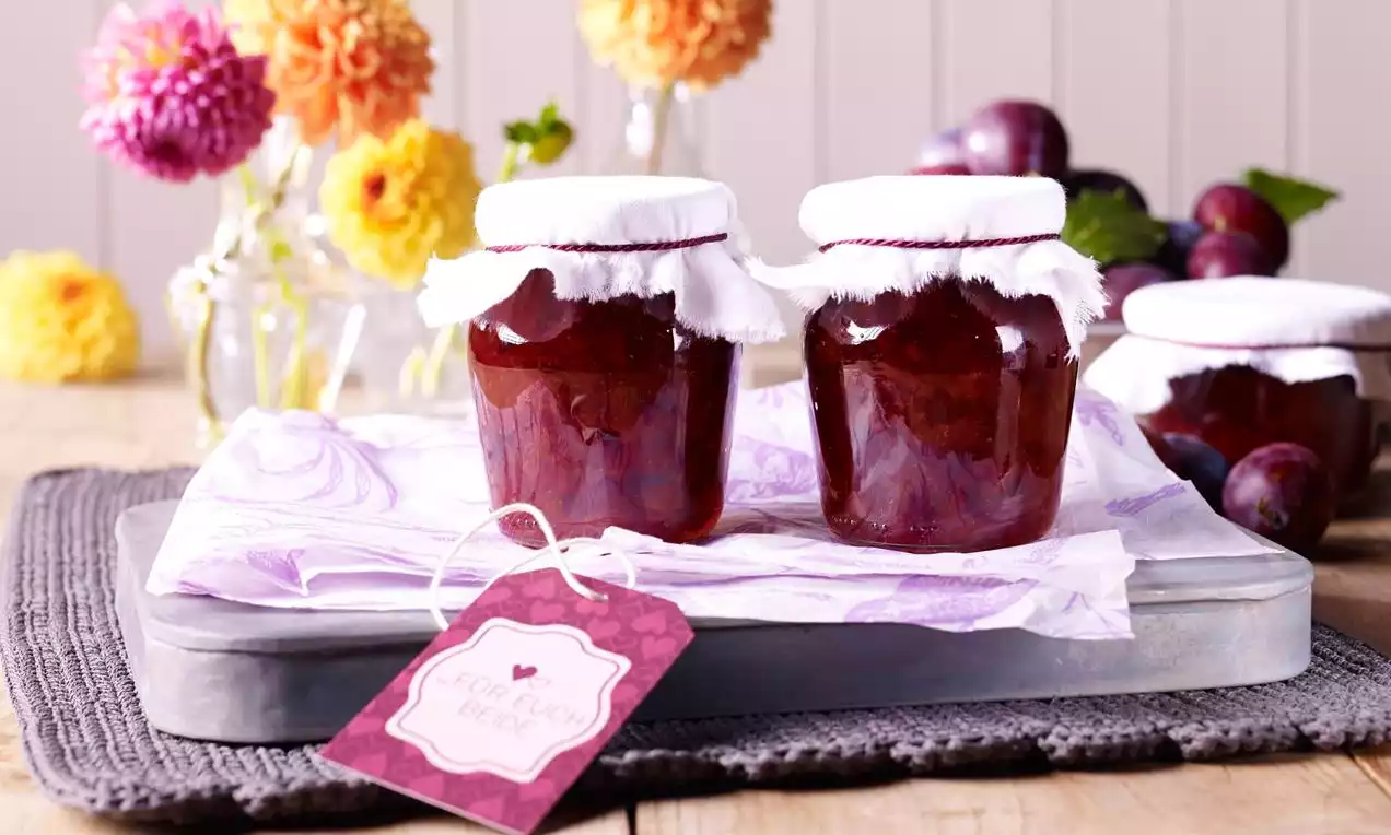 Rotweinpflaumenkonfitüre mit sattem dunkelrotem Glanz in Gläsern mit weißem Stoffdeckel auf Tablett, umgeben von Herbstblumen und Pflaumen