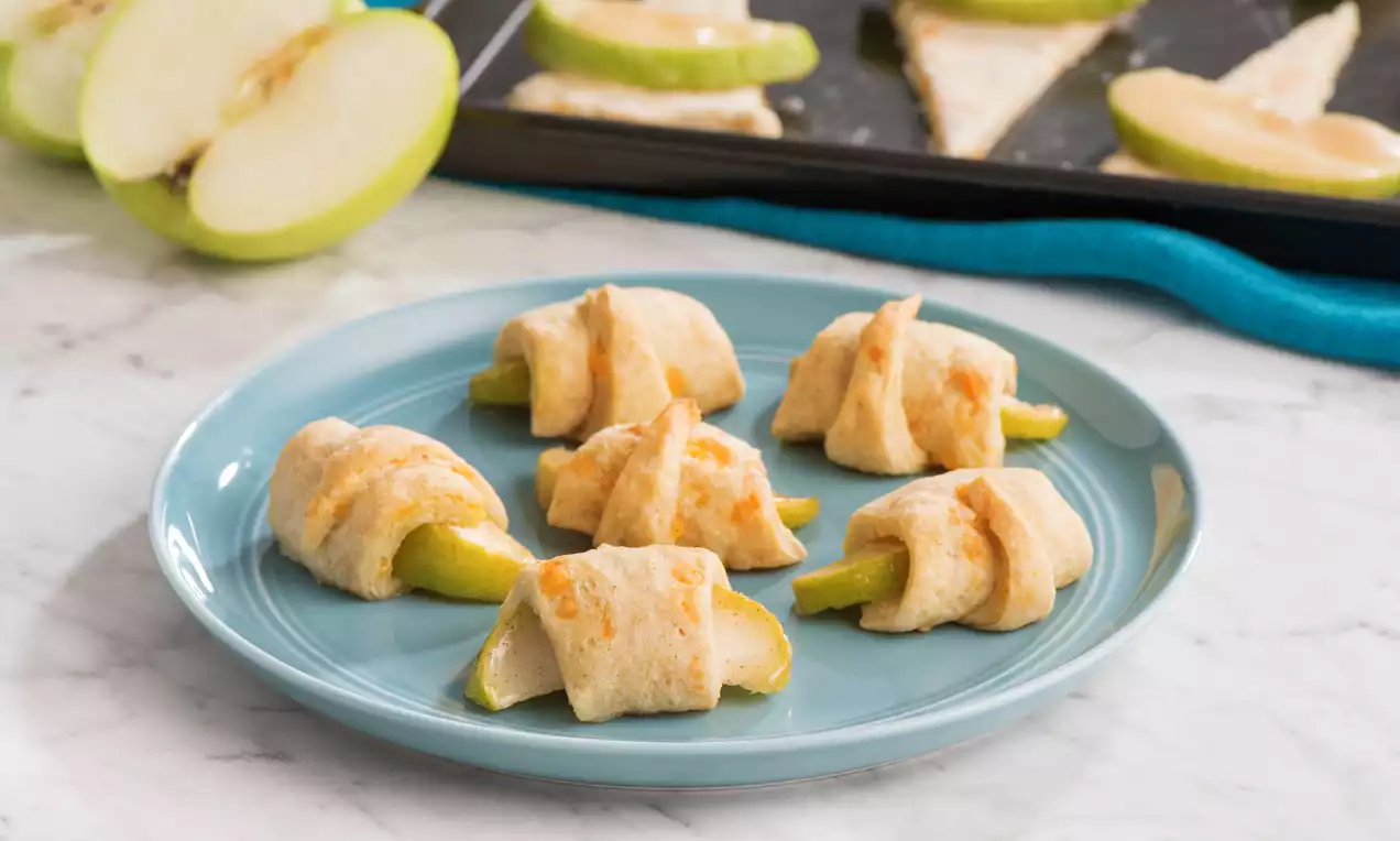 Golden crescent-shaped bites wrapped around vibrant green apple slices on a light blue plate, with a tray of apple pieces in the background