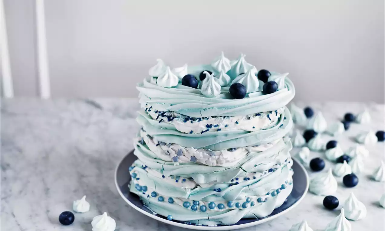 Høy lagdelt marengskake i lyseblå toner pyntet med blåbær, små marengstopper og perler på en mørk tallerken mot marmorbord