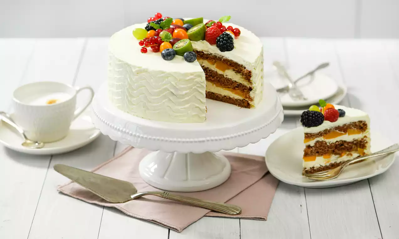 Mascarpone-Torte mit welliger weißer Creme, saftigem Schichtkuchen und buntem Obstbelag auf weißer Tortenplatte mit Kaffeegedeck