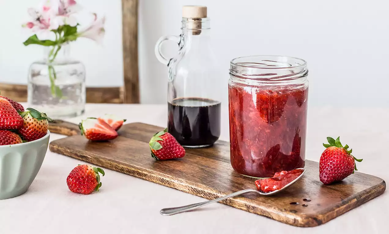 Sklenice jahodového džemu s hlubokě červenou barvou vedle lahvičky tmavého balsamika, čerstvých jahod a lžíce na dřevěné desce