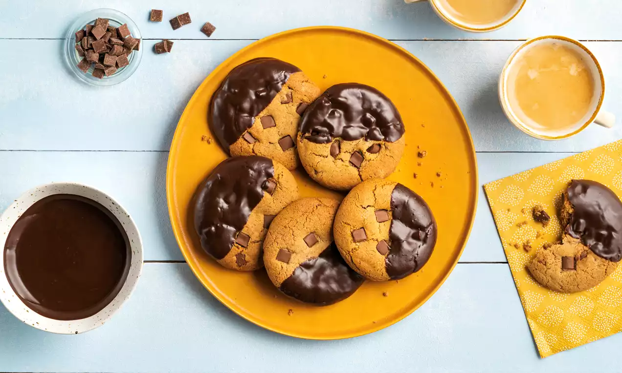 Ronde, goudbruine chocolate chip cookies met halve chocodip, gegarneerd met chocoladestukjes, op een geel bord met kopjes koffie