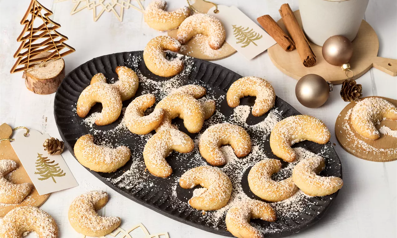 Goldbraune Zimtkipferl, leicht gebogen und mit Puderzucker bestäubt, auf schwarzer Platte, umgeben von weihnachtlicher Dekoration