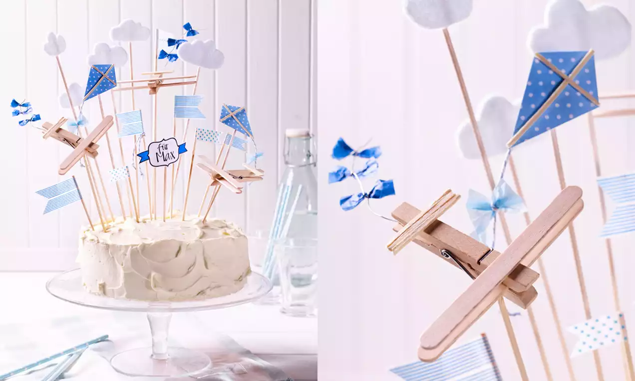 Vanille-Daphne-Torte mit heller, cremiger Oberfläche auf Glasplatte, dekoriert mit Holzflugzeugen, Wolken und blau-weiß karierten Papierdrachen