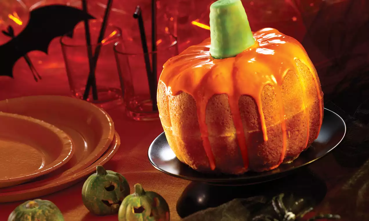 Gâteau en forme de citrouille, glacé orange vif avec une tige verte, posé sur une assiette noire, entouré de décoration d'Halloween
