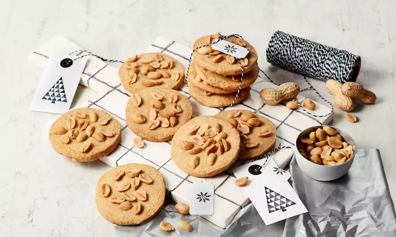 Goldbraune Erdnussplätzchen mit gerösteten Erdnüssen, flach und rund, auf einem karierten Tuch, daneben Nüsse und kleine Geschenkanhänger