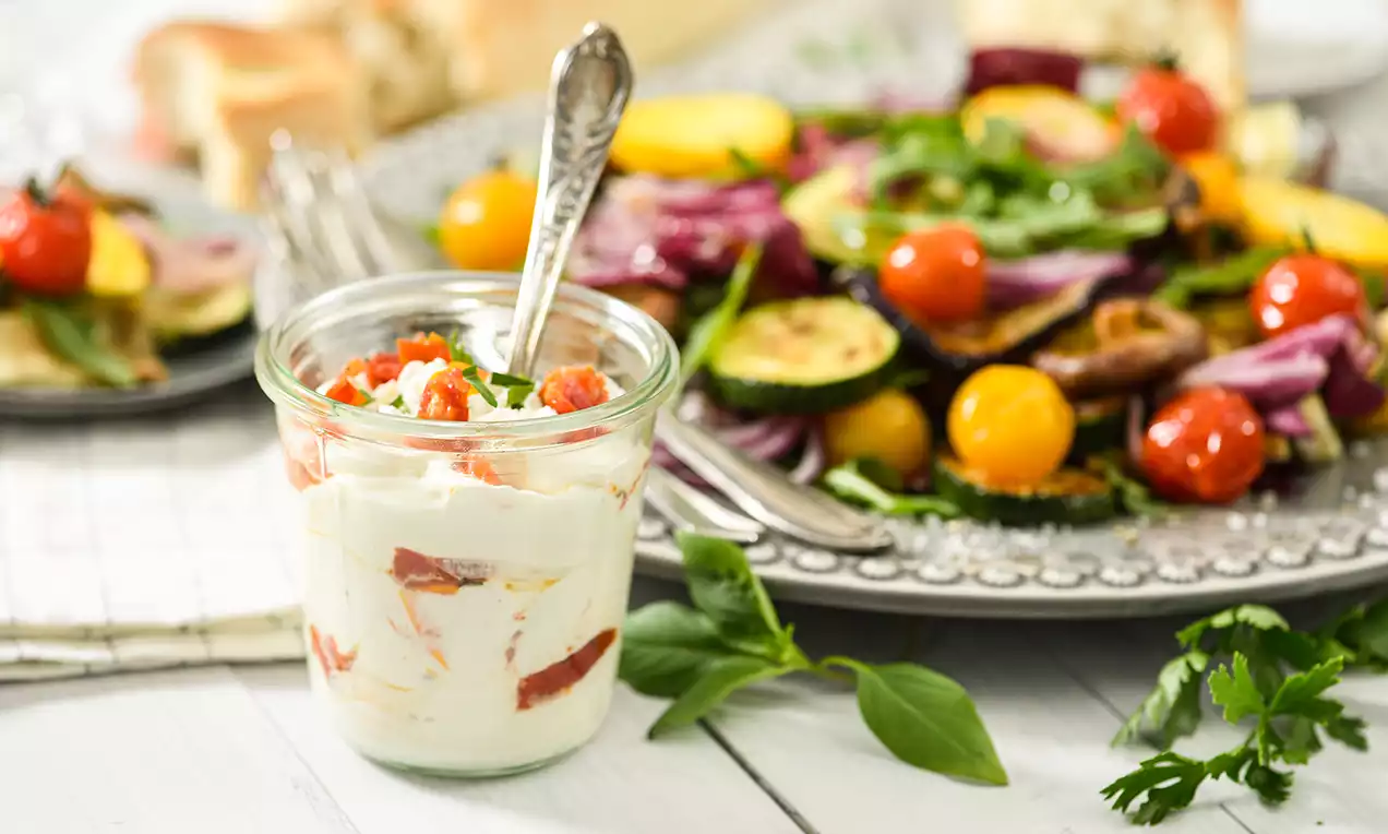 Crème-fraîche-Dip mit weißer, cremiger Textur und roten Tomatenstücken in einem Glas, garniert mit grünen Kräutern, daneben ein Teller mit Gemüse