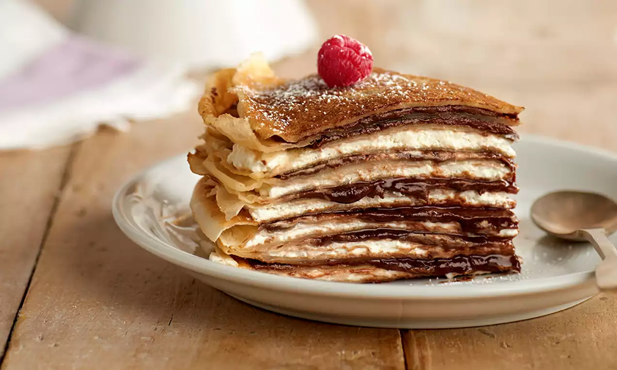 Lagen van goudbruine pannenkoeken met romige slagroom en glanzende chocoladevulling, gegarneerd met een framboos en poedersuiker op een wit bord