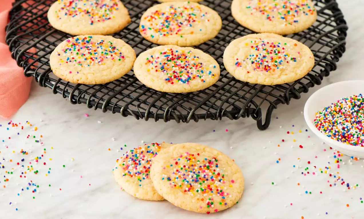 Cookies dorés et moelleux, parsemés de billes multicolores sur une grille noire avec une coupelle de vermicelles sur une table en marbre