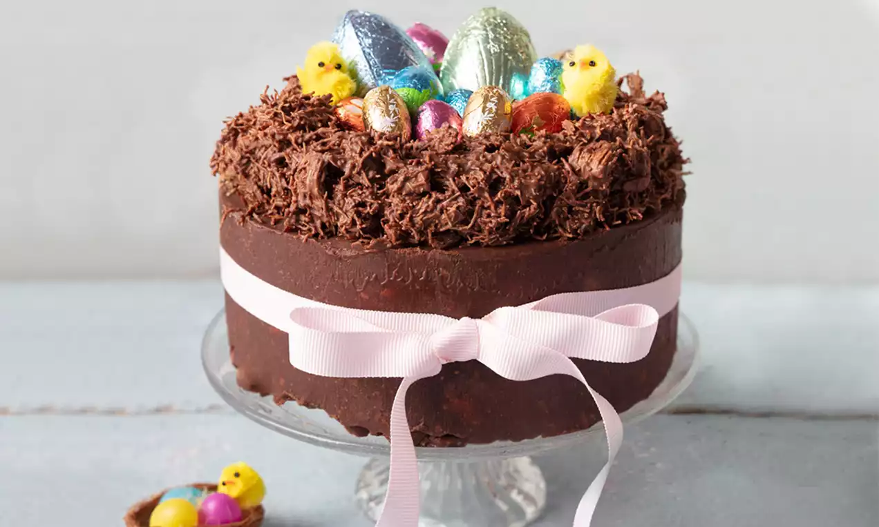 Tort rotund cu glazură lucioasă de ciocolată îmbrăcat într-o fundă roz, decorat cu un cuib din fulgi de ciocolată și ouă colorate