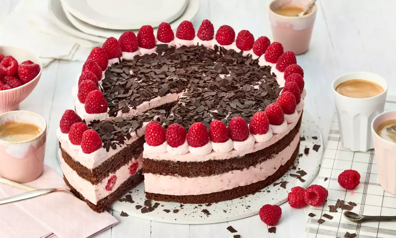 Schoko-Himbeer-Torte mit zwei dunklen Böden, rosa Creme und Himbeeren, mit Schokoraspeln bestreut auf weißem Teller mit Kaffee und Himbeeren