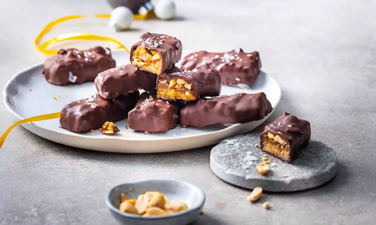 Chokladtäckta rektangulära bars fyllda med kola och rostade jordnötter, arrangerade på ett fat med grå stenplatta och lösa jordnötter