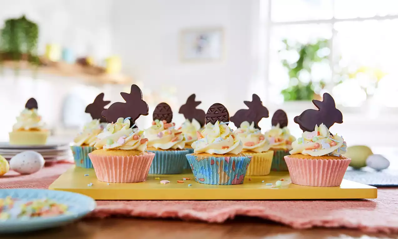 Easter cupcakes in pastel cases topped with swirls of cream frosting, chocolate bunny and egg decorations, and colourful sprinkles on a yellow board