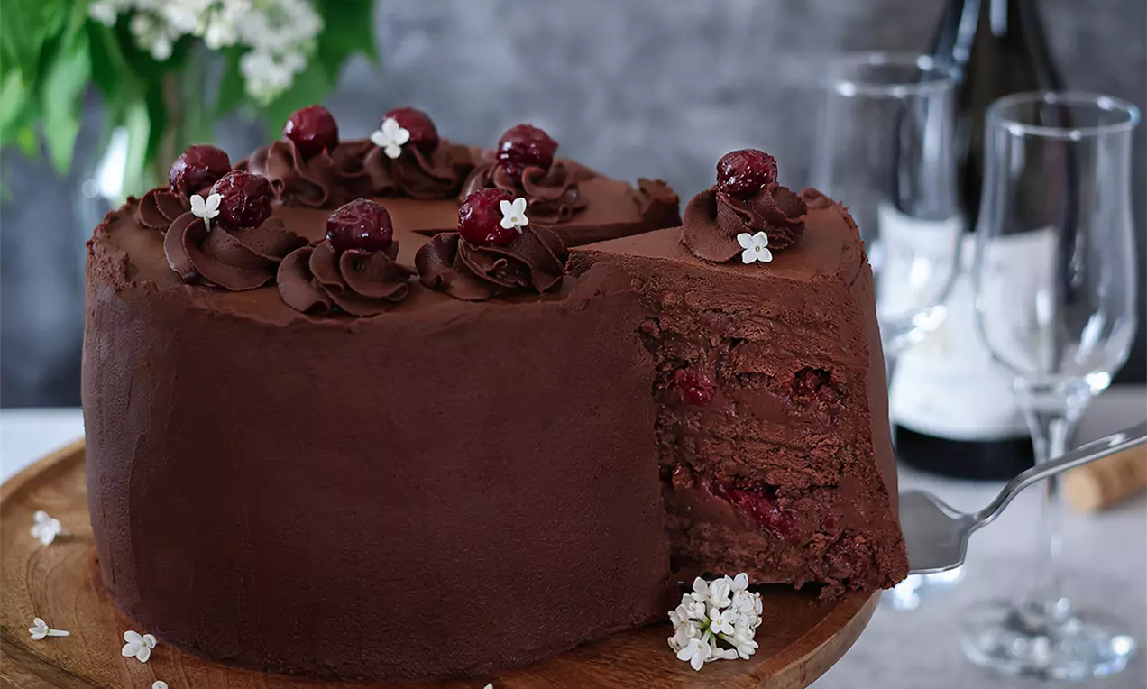 Шоколадова бисквитена торта с наситено кафяв крем, гарнирана с вишни и малки бели цветчета върху дървена подложка, с изрязан парче