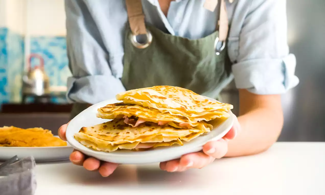 Crepes dorados y finos doblados en triángulos, con jamón y queso visibles en capas, servidos sobre un plato blanco sostenido por manos