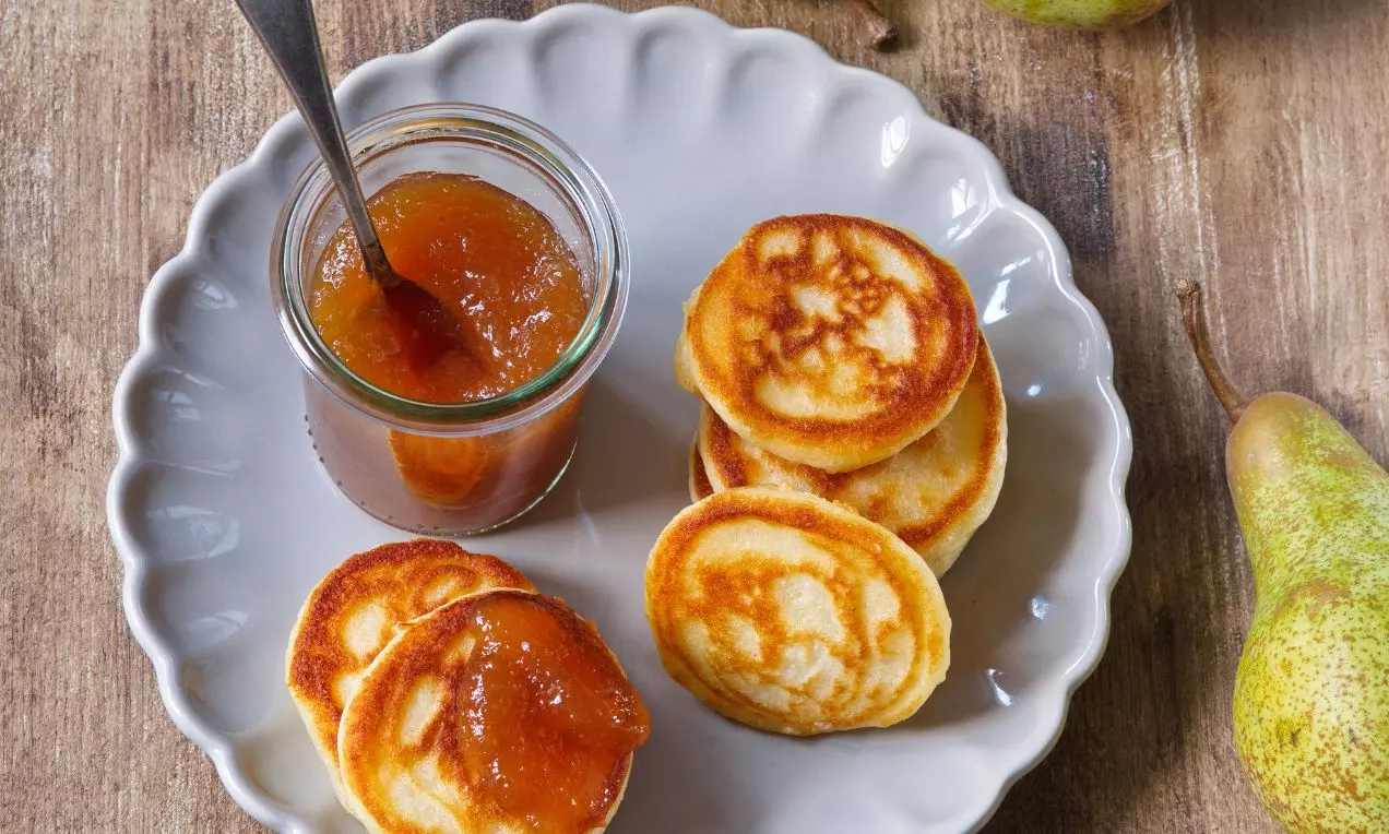 Hruškový džem jantárovej farby v skle so striebornou lyžičkou, vedľa zlatistých lievancov na zvlnenom bielom tanieri, doplnené čerstvou hruškou