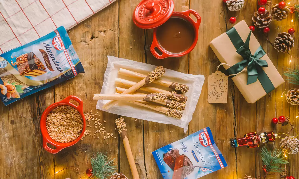 Neulas doradas y alargadas con extremos cubiertos de chocolate y crocanti, dispuestas sobre papel blanco junto a adornos navideños