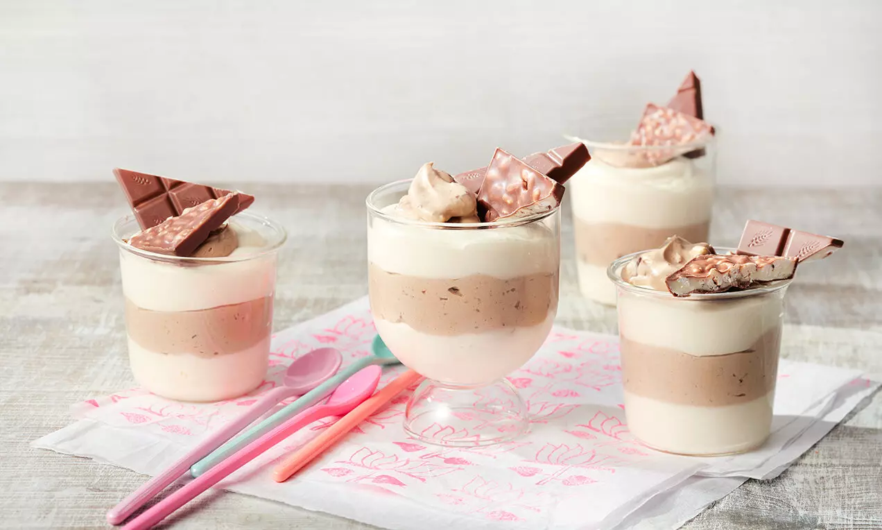 Слоеве от нежен крем в чаши със светло кафяв и бял цвят, завършени с шоколадови триъгълници върху розова салфетка