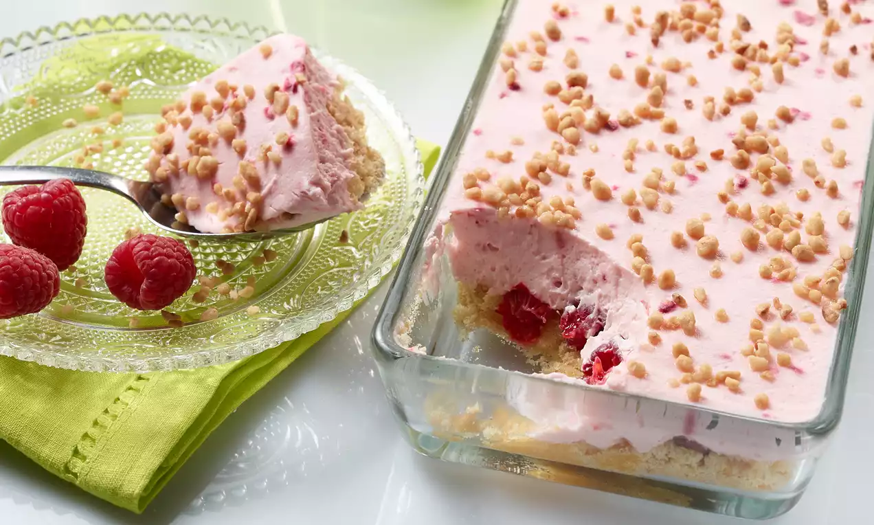 Tiramisu slojevit s ružičastim kremom, zlatnosmeđim posipom i crvenim jagodama u staklenoj posudi, uz tanjir s malinama na zelenom ubrusu