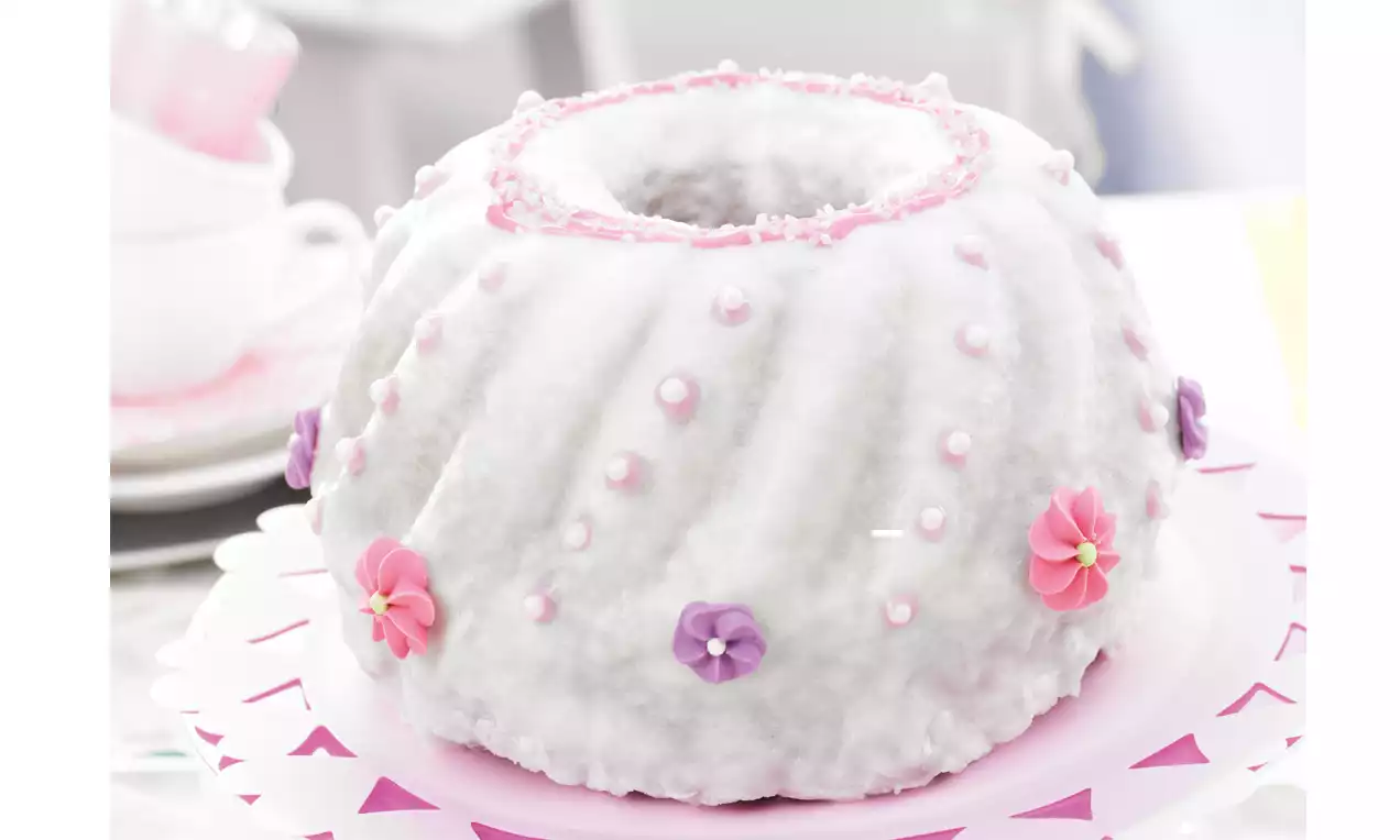 Ein weiß glasierter Gugelhupf mit rosa und lila Zuckerdekor, Perlen und Blüten auf einer pastellrosa Tortenplatte mit Zickzackmuster