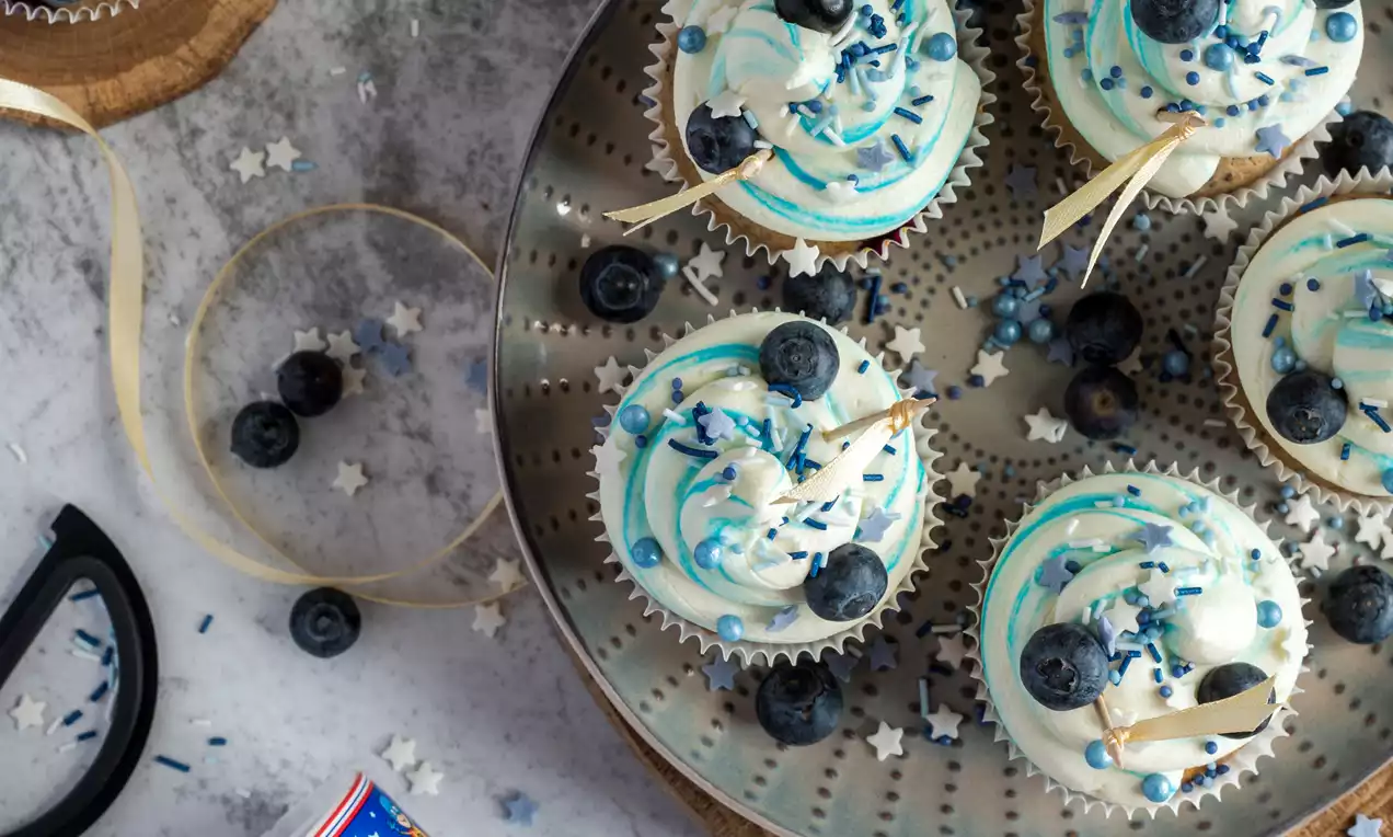 Citrondzelteni keksiņi ar krēmīgas baltas putukrējuma virpuļiem, rotāti ar tumšām melleņu ogām un ziliem cukura rotājumiem uz metāla šķīvja