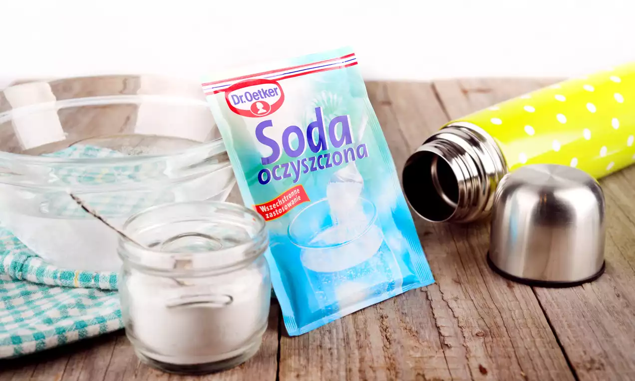 Przezroczysta miska z wodą na drewnianym blacie obok słoika z białym proszkiem, zielonego termosu w groszki i opakowania sody oczyszczonej