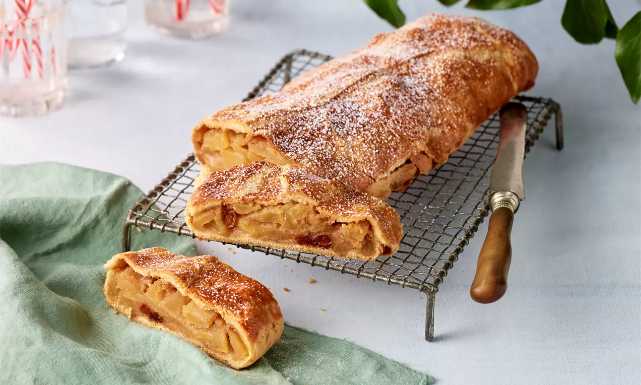 Goldbrauner Apfelstrudel aus Mürbteig mit sichtbarer Füllung, bestäubt mit Puderzucker auf einem Gitterrost neben einem Messer und Stoffserviette