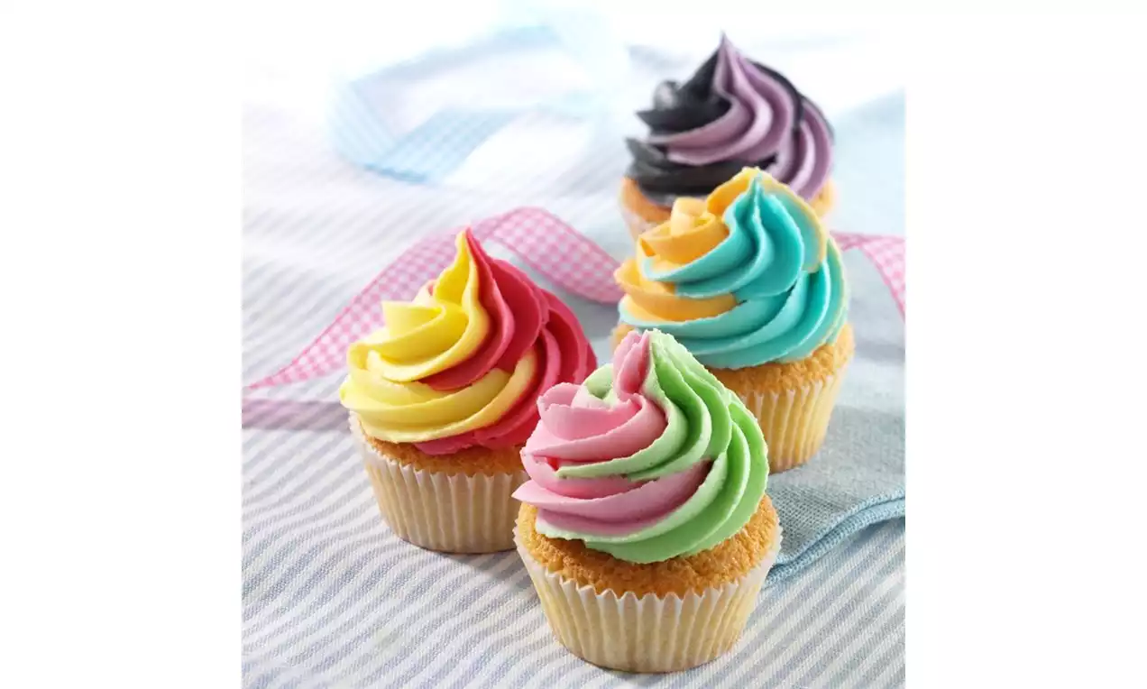 Four fairy cakes with golden bases topped with smooth two-tone swirls in pink, yellow, green, blue, and purple set on striped fabric