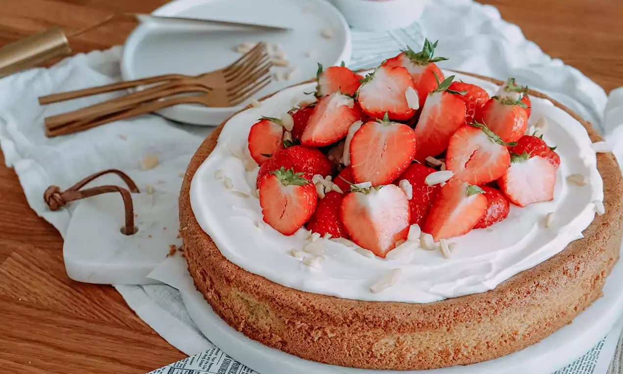 Goldbrauner Tortenboden mit weißer Creme, darauf ganze frische Erdbeeren und Streusel, drapiert auf einer weißen Platte mit Besteck im Hintergrund