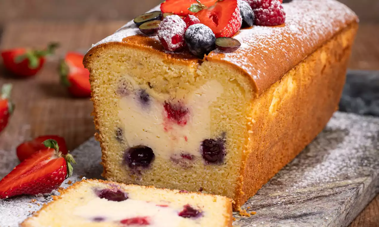 Pravokotni cheesecake kolač z mehko vaniljevo sredico in jagodičevjem, prelit z jagodnim prelivom in posut s sladkorjem v prahu na leseni deski