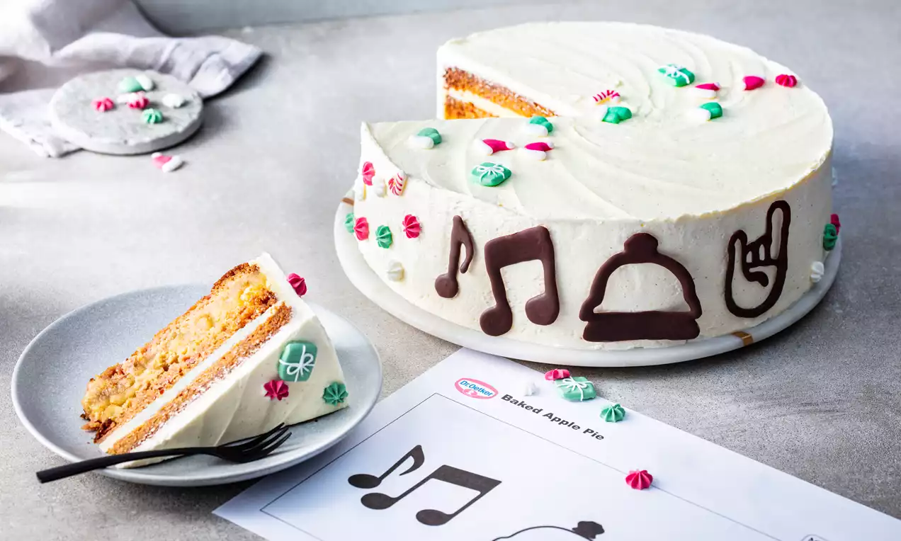 Gâteau blanc recouvert de crème lisse, décoré de symboles en chocolat noir et de petites rosettes colorées, avec une part coupée sur une assiette grise.