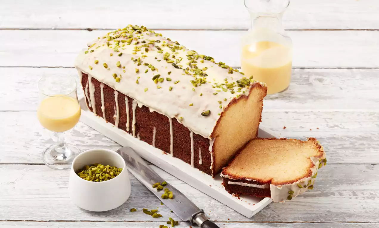 Kastenkuchen mit goldgelbem Teig, weißer Glasur und gehackten grünen Pistazien, angerichtet auf weißer Platte mit Messer und Eierlikörglas