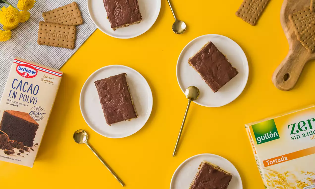 Porciones rectangulares de tarta con capas de galleta tostada y crema cubiertas con una capa lisa de chocolate en platos blancos sobre una mesa amarilla