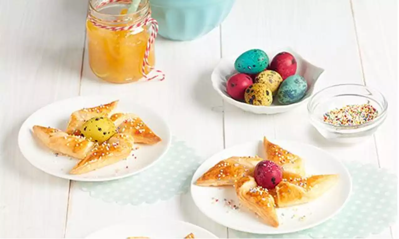 Molinillos de hojaldre dorado con mermelada vibrante al centro, espolvoreados con azúcar y grageas coloridas en platos blancos sobre mesa clara