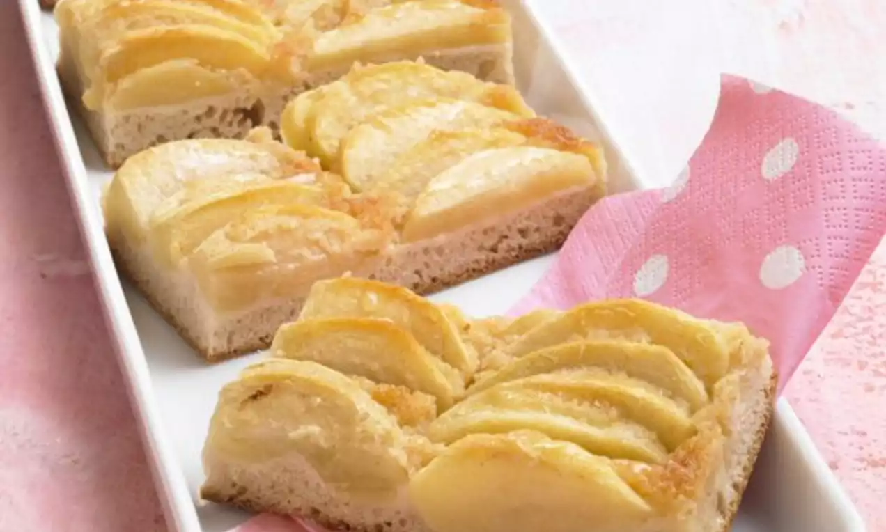 Tranches de pomme dorées et juteuses sur une pâte moelleuse aux bords légèrement croustillants, servies dans un plat blanc avec une serviette rose à pois